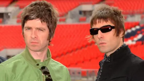 PA Media Noel e Liam Gallagher franzindo a testa para a câmera em uma foto tirada em 2008. Noel está vestindo uma jaqueta verde e Liam uma preta. Ele está usando óculos escuros. Ambos têm penteados modernos.