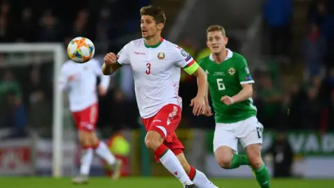 Getty Images Um jogador de futebol bielorrusso, vestindo uma camisa branca, shorts vermelhos e meias brancas, persegue uma bola de futebol. Ele é perseguido por um jogador da Irlanda do Norte de camisa verde, calção branco e meias verdes