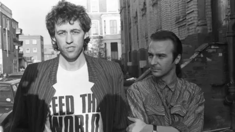 Getty Images Uma foto de arquivo de Bob Geldof vestindo uma camiseta que diz 'alimente o mundo' ao lado de Midge Ure.