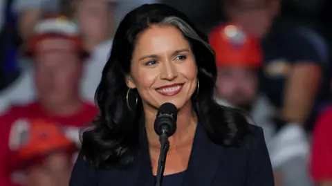 Reuters A ex-deputada norte-americana Tulsi Gabbard participa de um comício de campanha do candidato presidencial republicano e ex-presidente dos EUA, Donald Trump, na PPG Paints Arena em Pittsburgh, Pensilvânia - ela está diante do microfone, tem um sorriso largo e brincos de ouro com uma jaqueta escura e principal. Ela tem uma mecha branca distinta no cabelo e uma multidão borrada pode ser vista atrás dela.