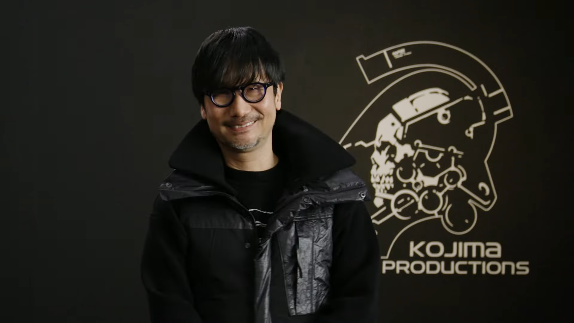 Uma foto mostrando um Hideo Kojima sorridente em frente ao logotipo da Kojima Productions após a revelação do Physint