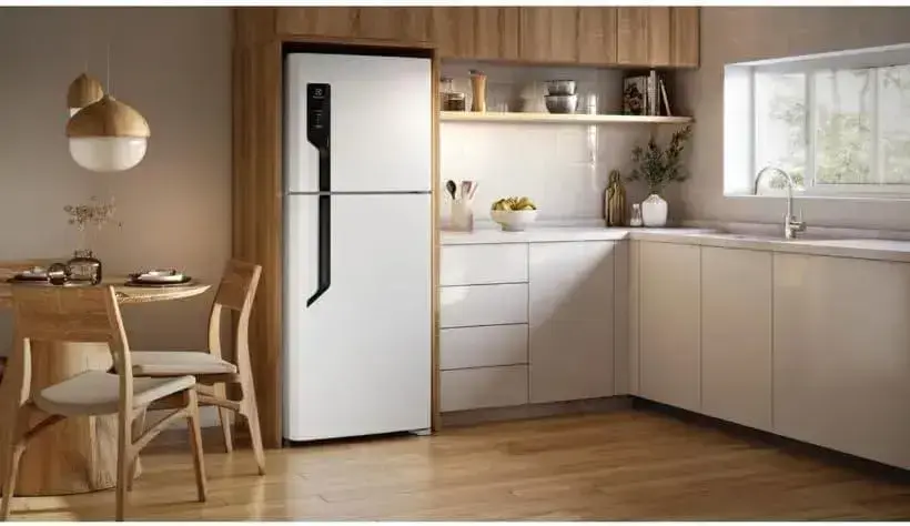 Refrigerador frontal na cozinha com paredes em tom claro, prateleira de madeira e iluminação natural.